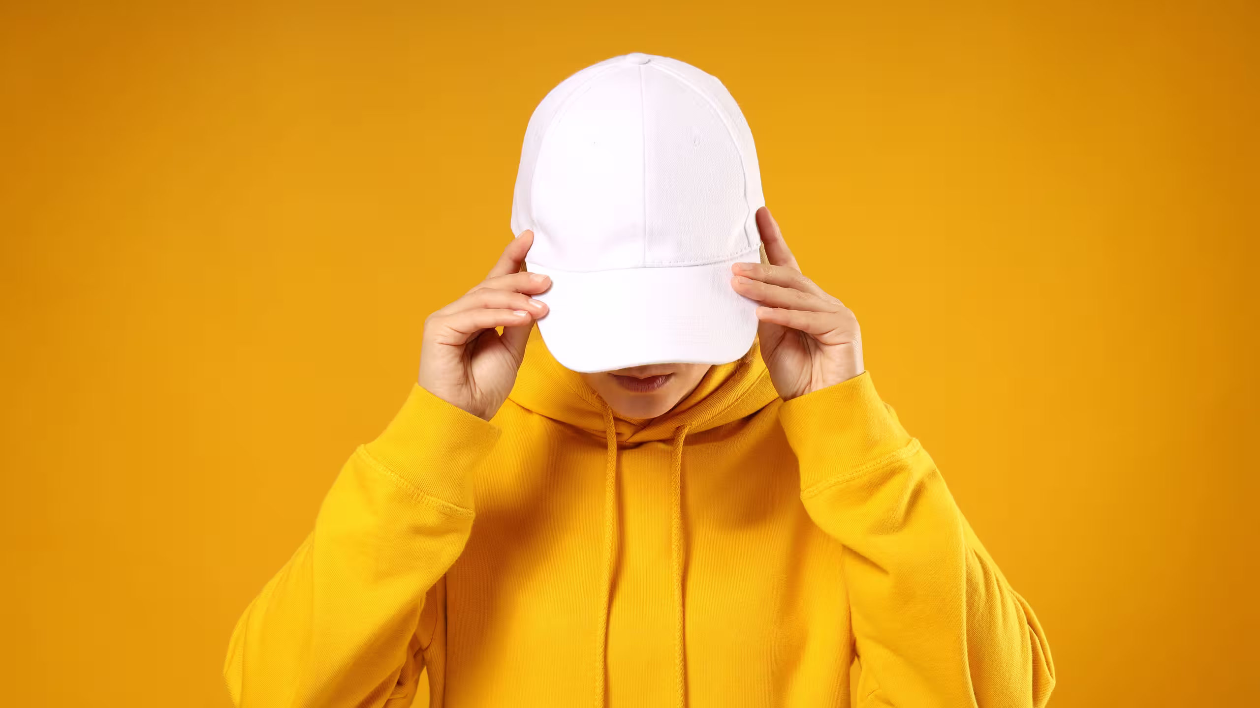Person in a yellow hoodie adjusting a white baseball cap, set against a solid orange background.